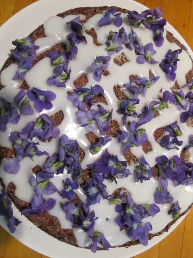 a single layer cake topped with a drizzled vanilla glaze and fresh violet flowers.