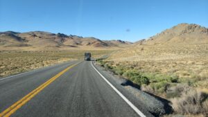 A photo taken through the windshield while driving on a two-lane winding highway through rolling desert hills with brown and green brushy low foliage. A RV is on the road ahead. 
