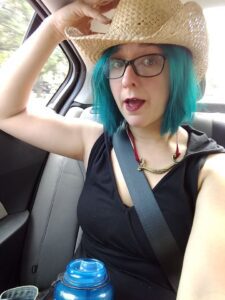 A girl with turquoise hair, glasses, dark red lipstick, wearing a black dress, a brass and velvet necklace and a straw cowboy hat sits in the backseat of a car holding a water bottle.
