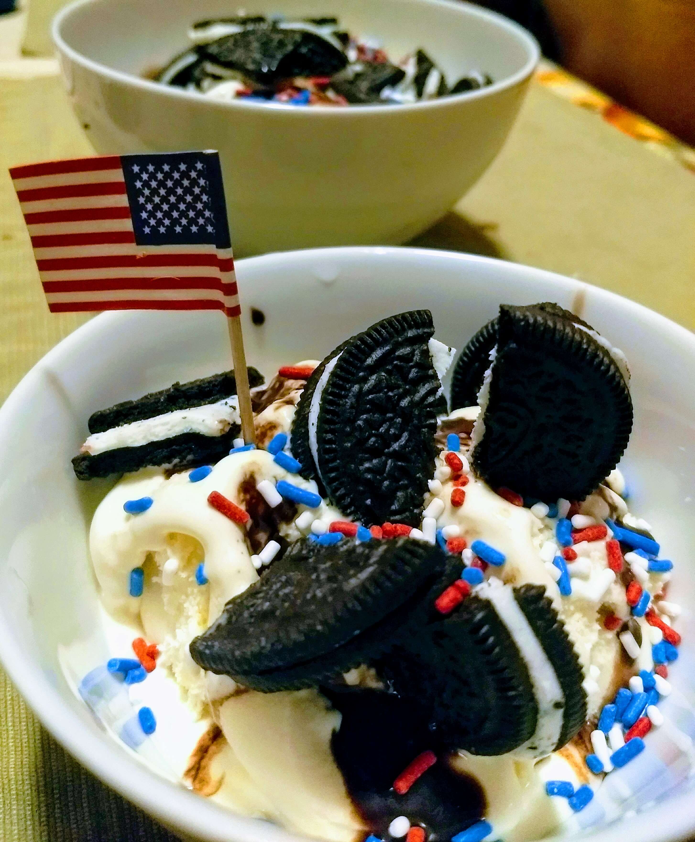 an ice cream sundae with crushed oreos, vanilla ice cream, red white and blue sprinkles and an American flag toothpick decor