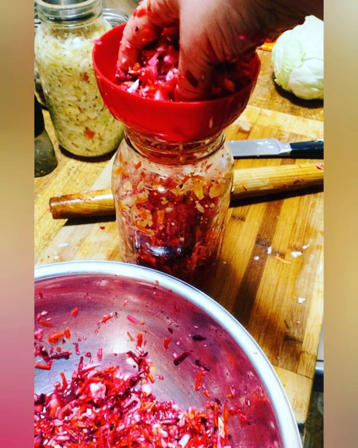 a bowl of shredded vegetables being packed carefully into a mason jar using a canning jar funnel and a wooden rolling pin to make homemade sauerkraut