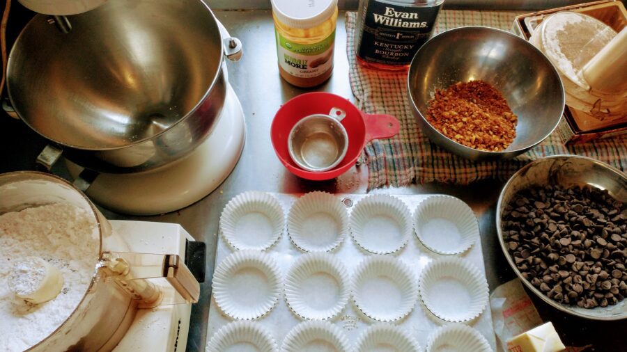mise en place laid out for homemade peanut butter cups, with paper lined muffin tins, a food processor, peanut butter, bourbon, crushed peanut brittle, and chocolate chips on a kitchen counter.