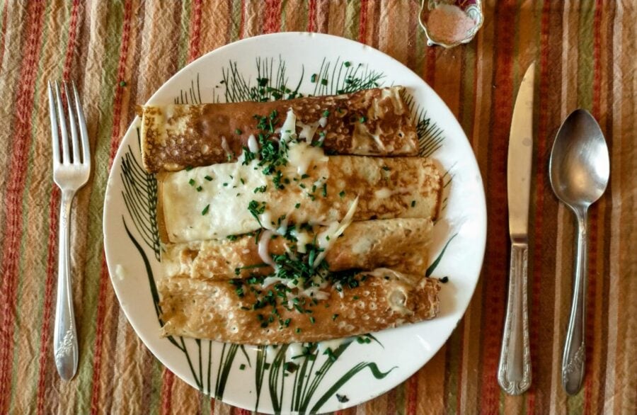 spinach artichoke crepe recipe. Image of a semi-formal table setting with vintage silverware, a striped orange and green placemat, and a small gilded salt bowl, with a plate of filled, rolled crepes in the center.