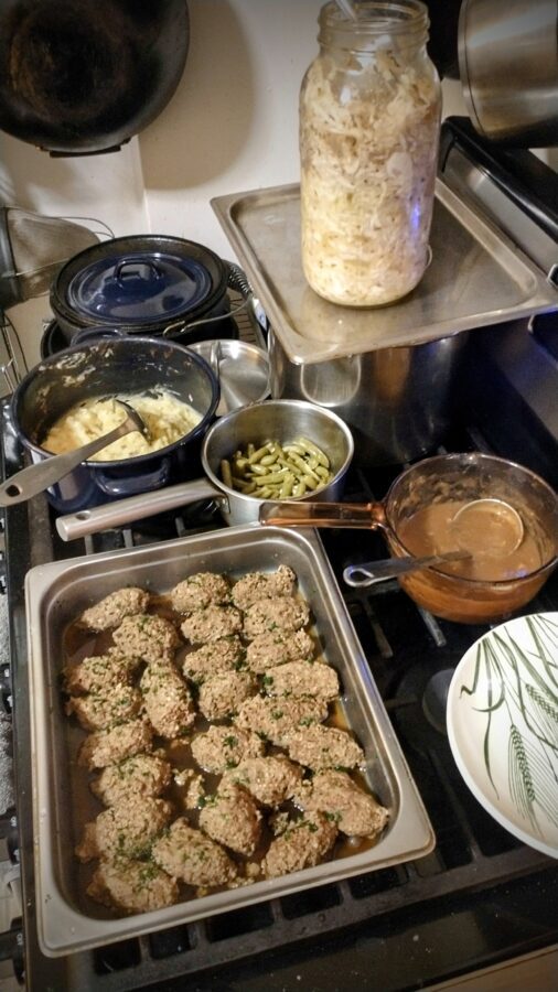 a stovetop with poached liver dumplings, mashed potatoes, green beans, gravy, and a jar of homemade kraut ready to serve.