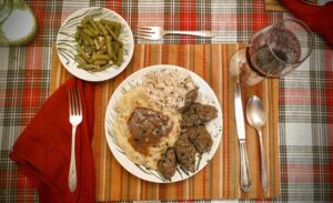 place setting with a side dish of green beans with bacon, mashed potatoes and mushroom gravy, sauerkraut, and German liver dumplings with a glass of red wine on a striped placemat