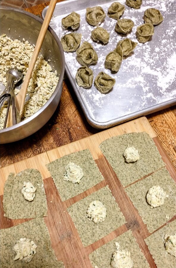 a bowl of tortellini filling, a cutting board with squares of green pasta dough and a dollop of filling, and a sheet pan with filled and shaped tortellini
