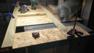 Boards, tools, and a roll of hardware cloth are laid out on a workbench for building a chicken brooder