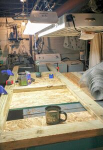 The chicken coop brooder frame is taking shape, clamped to the workbench and getting screwed together. A big mug of coffee is also on the bench. A laundry room area is in the background