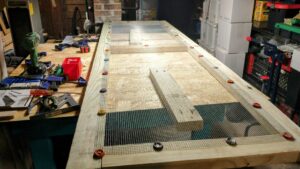 a side panel for a chicken brooder and grow out pen sits on a workbench. It's made from 2x4 lumber with wire mesh hardware cloth secured with bottle cap washers. An array of tools are visible, including Kreg jigs for pocket holes, clamps, and cordless Metabo drills