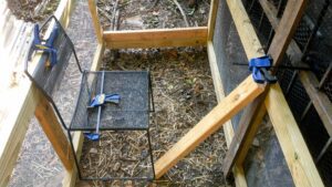 the fully assembled and hardware cloth covered frames are outside in the chicken run. They're being connected by other boards to make a rectangular frame. The sides are being supported by a fence, a chair, and clamps while they're being screwed together.