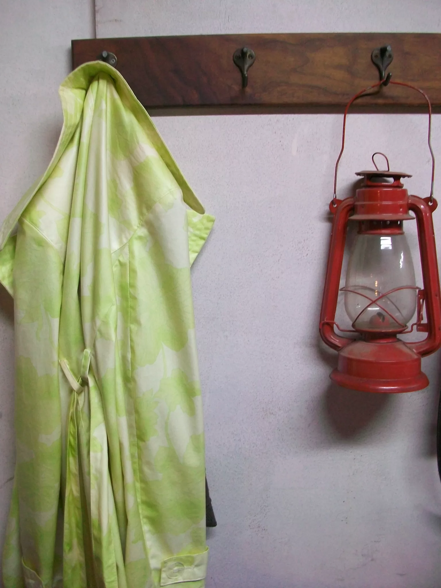A bright green and white floral print raincoat hangs on a hook next to a red kerosene lantern