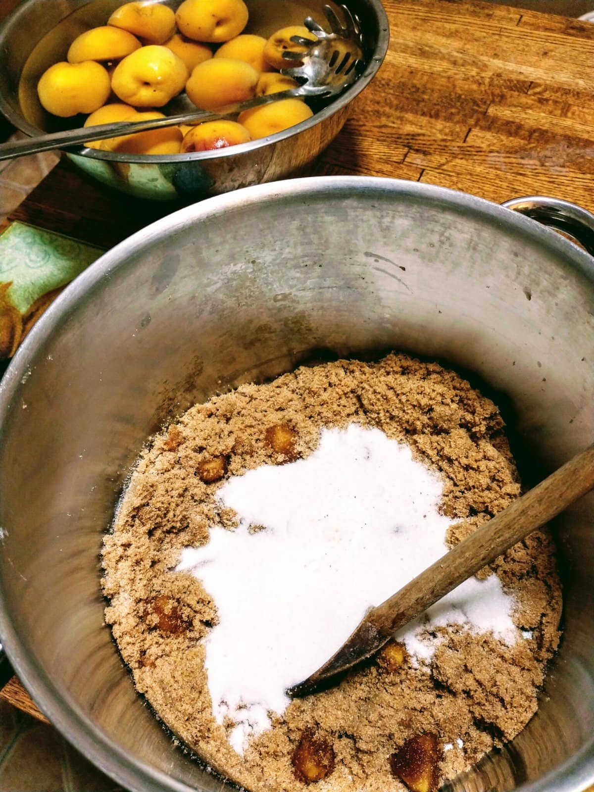 blanched fresh peaches float in a bowl of ice water, with a pot of brown and white sugar in the foreground. 