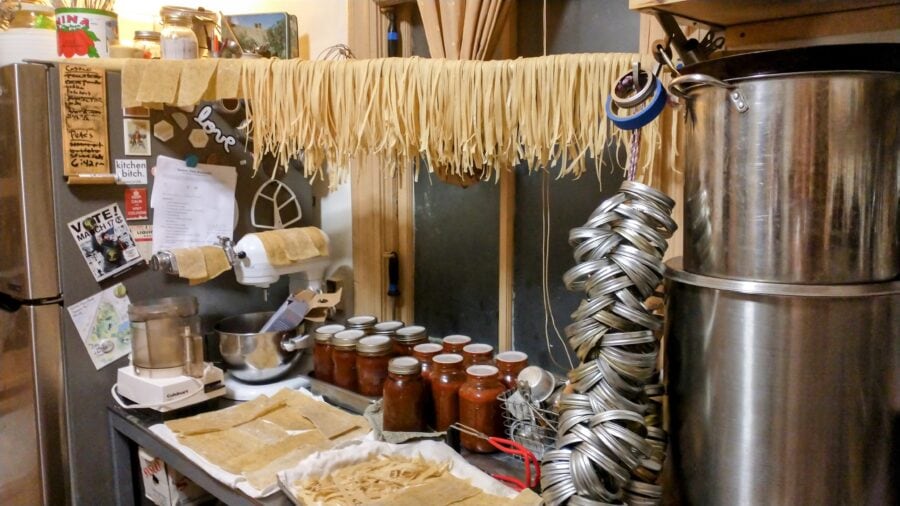 Fresh homemade pasta (egg noodle fettuccine) is drying on a bamboo pole hung between two shelves in a kitchen. Large pots and canning jar rings are in the foreground, and in the background trays of homemade pasta are drying and canned mason jars full of tomato ragu are on the counter