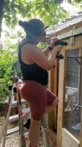 The author stands on a short ladder outside in a garden with lots of trees. Her hair is in a bun and she's wearing a tank top and bike shorts. She is using a drill to make pilot holes to install barn-door hardware on a chicken coop door.