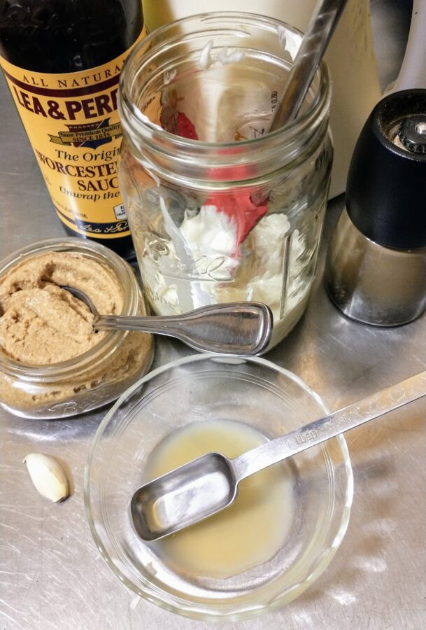 Photo shows bottles and jars of ingredients (lemon juice, worstershire sauce, dijon mustard, mayonnaise, a garlic clove, and a black pepper grinder)