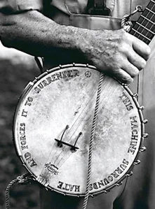 a black and white close-up photo of Pete Seeger, holding his weathered banjo. The banjo has writing on the face- "this machine surrounds hate and forces it to surrender"