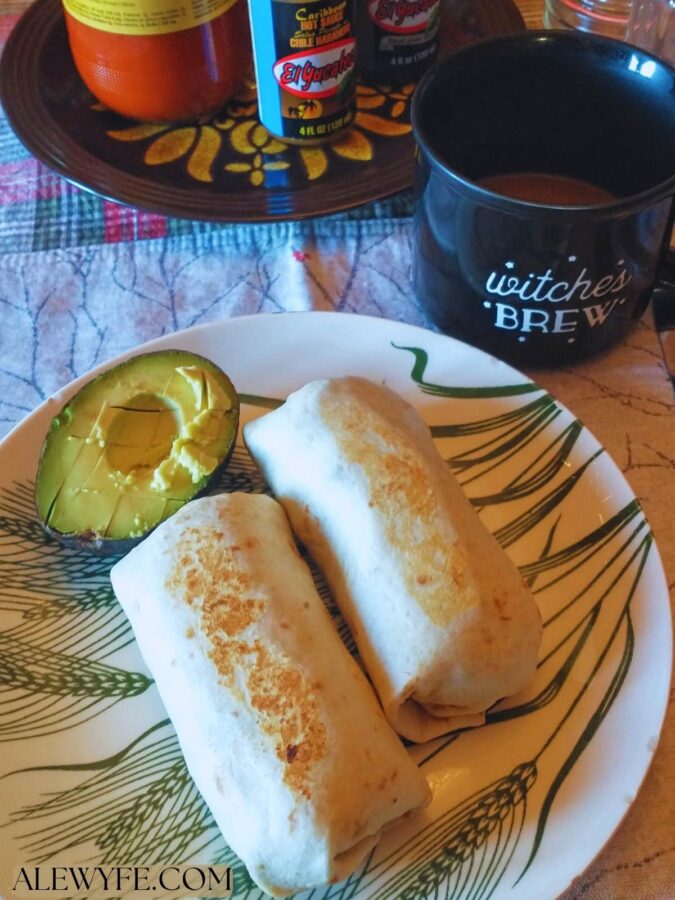 two small burritos on plate with half an avocado, a mug, and hot sauces