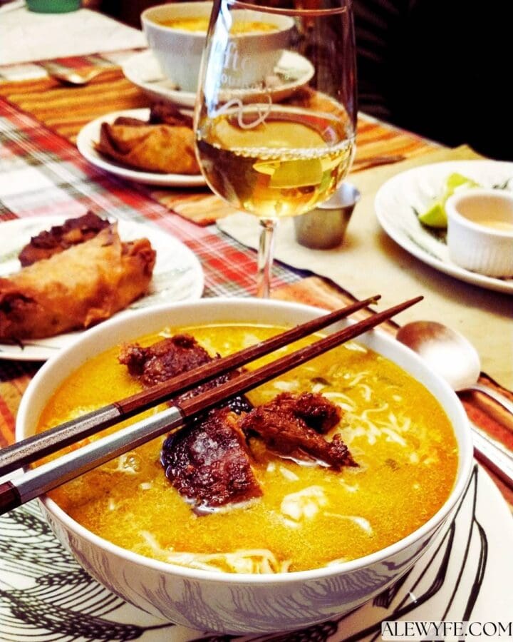 a white bowl of ramen with crispy tofu and bamboo chopsticks. a glass of white wine in stemware, and side plates with an egg roll and pot sticker. 