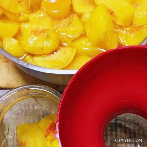 a bowl of summer peach slices being packed into jars using a bright red canning funnel