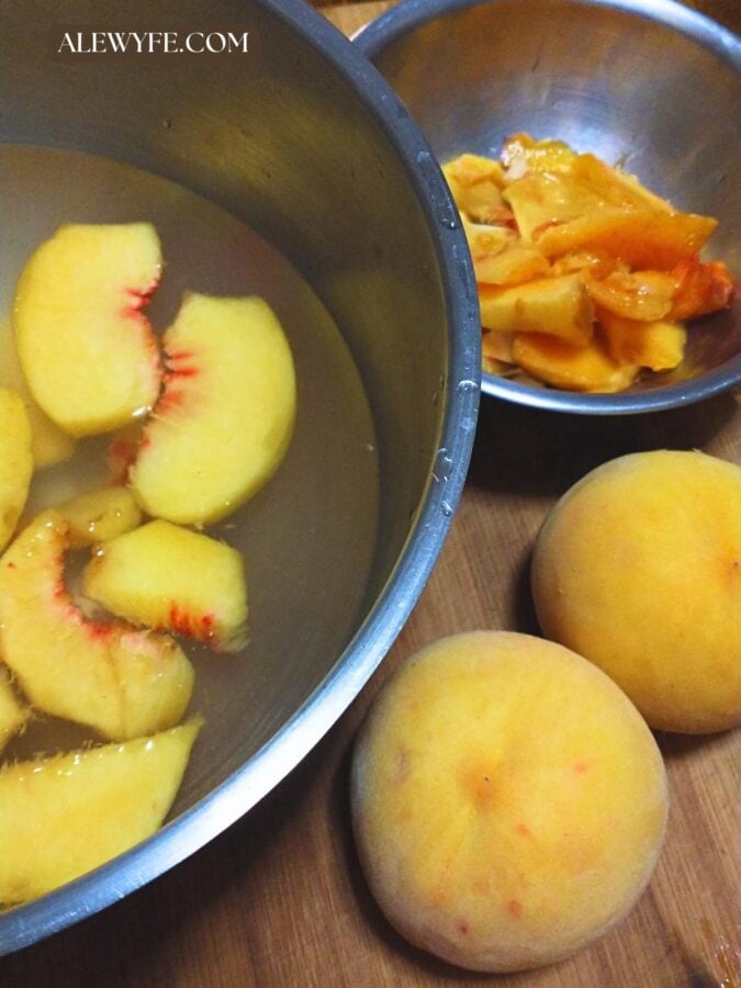 a stainless steel mixing bowl of peach slices floating in lemon water along with two whole peaches and a bowl of trimmings for peach nectar