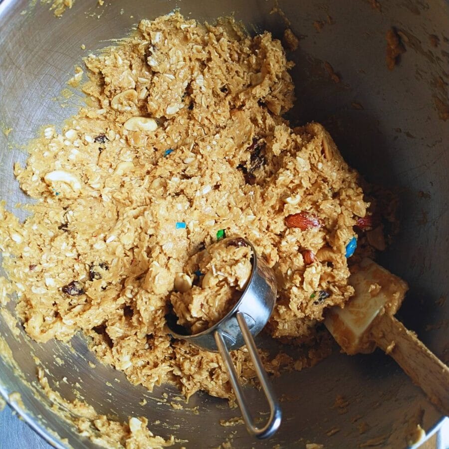 mixing bowl with dough for peanut butter oatmeal trail mix cookies with a half-cup metal scoop and a spatula.