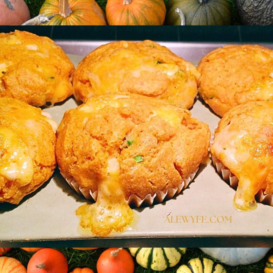 a pan of savory pumpkin muffins in the oven, with melted cheese oozing out as they bake. there is a background of colorful pumpkins and fall gourds framing the photo.