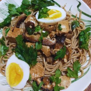 Close up photo of a bowl of cold soba noodles with a halved jammy egg, sliced shitake mushrooms, marinated tofu, and cilantro leaves