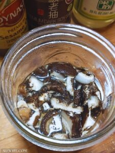 sliced shitake mushrooms rehydrating in a mason jar, with bottles of sesame oil, dark soy sauce, and rice vinegar in the background to make cold soba noodle dressing