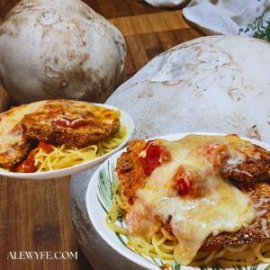 two wide bowls heaped with buttered spaghetti and topped with puffball parmigiana, with layers of red marinara sauce, fried puffball mushroom slices, and melted cheese. background image of two large puffball mushrooms on a cutting board