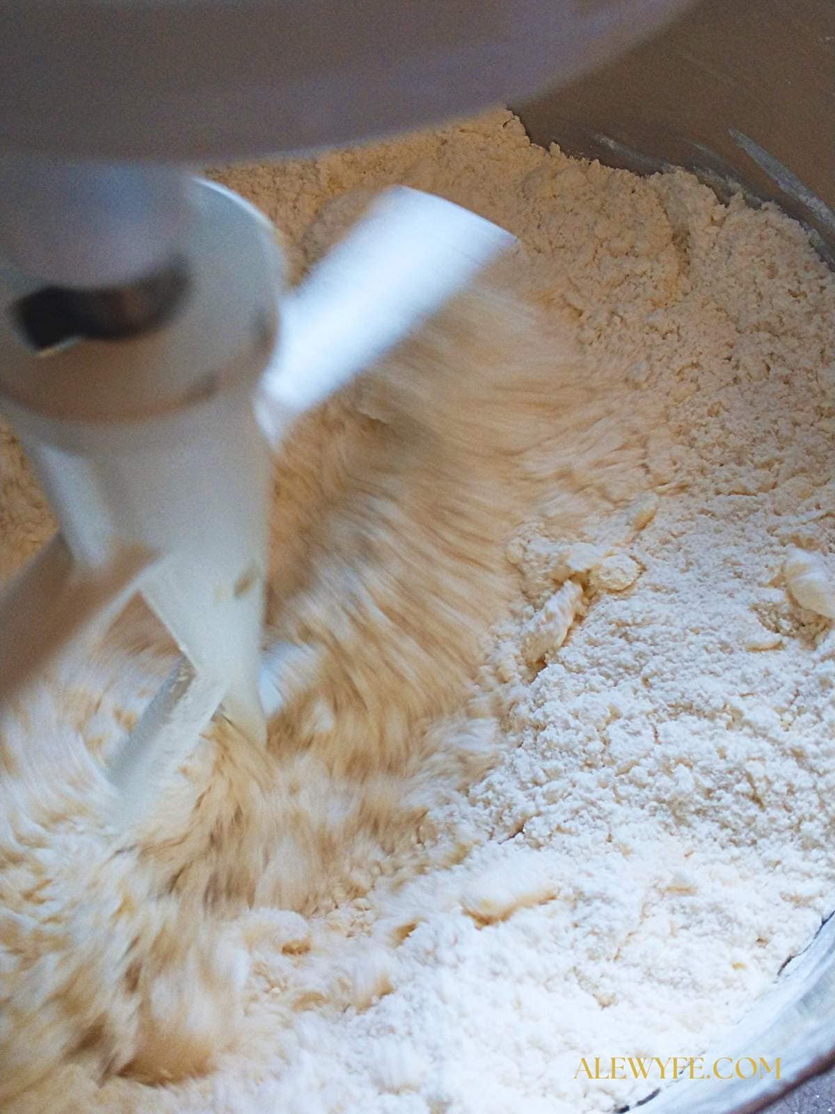 a stand mixer bowl filled with a crumbly mixture of butter, flour, and salt for pie crust dough. The mixer paddle attachment is blurred from mixing the dough