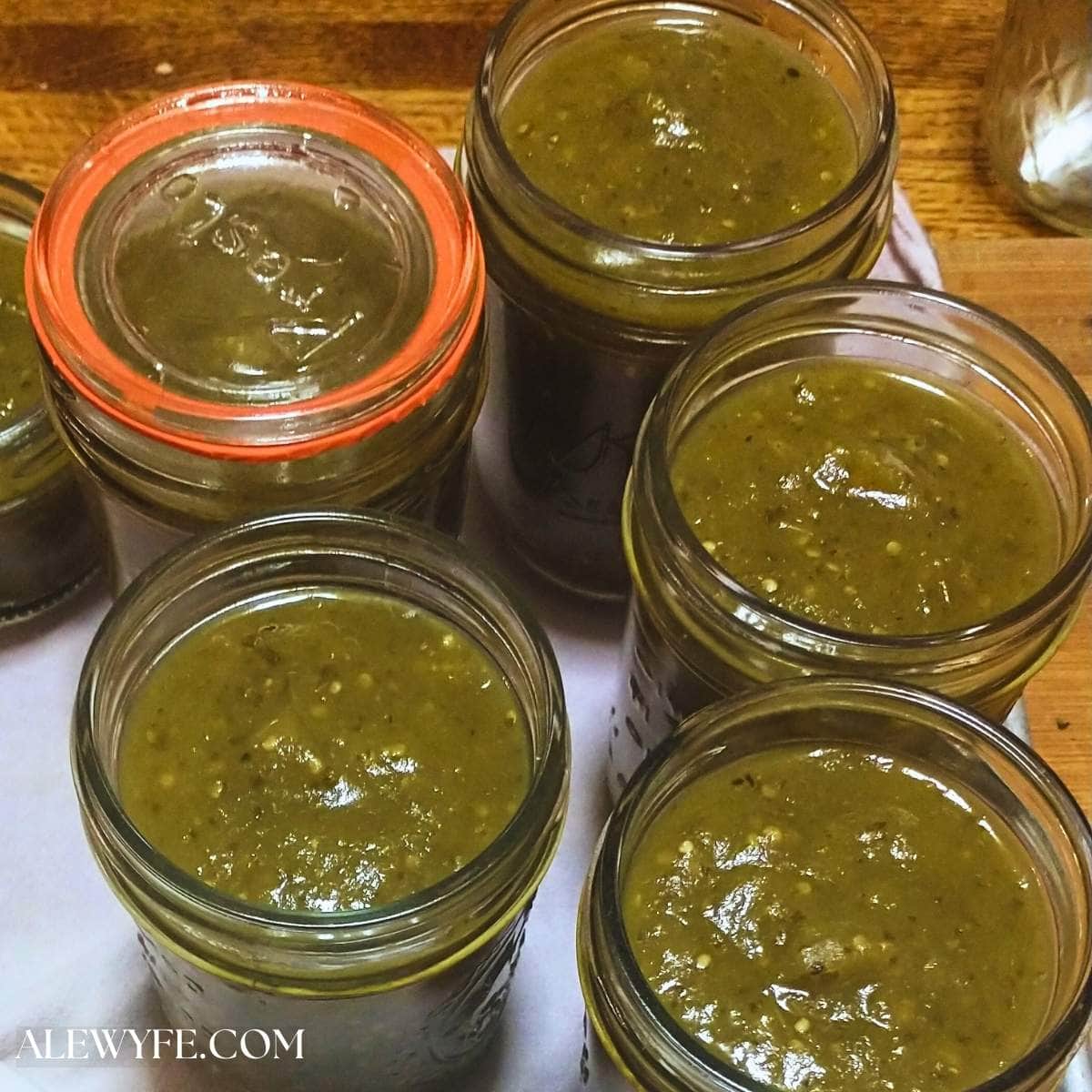 filled jars of roasted tomatillo salsa verde, ready for canning. one jar has an old-fashioned glass lid with orange rubber seal and the others are still uncovered