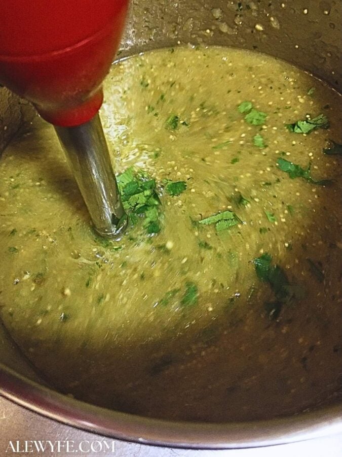 a red immersion blender in a pot of salsa verde, pureeing the roasted vegetables into a smooth salsa