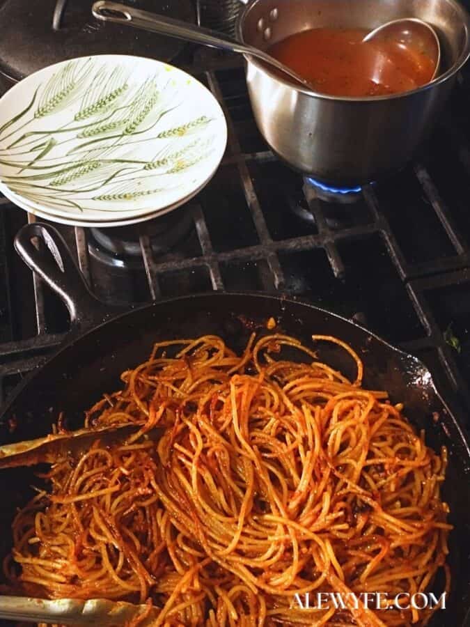 A cast iron pan with sizzling assassin's spaghetti cooks on a gas stovetop, with a pot of tomato broth and a ladle, and serving bowls at the ready