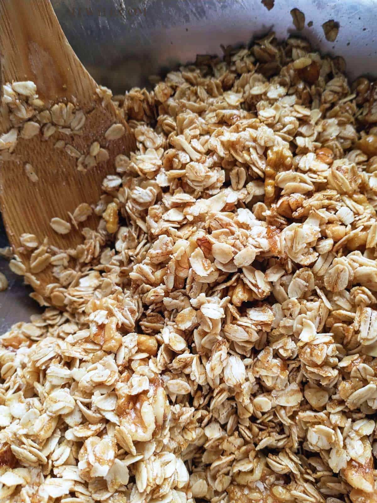 a stainless bowl filled with rolled oats, nuts, honey, and melted butter being stirred with a wide bamboo spoon