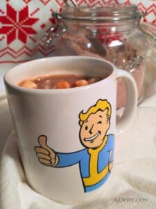 hot cocoa with marshmallows in a mug printed with Vault Boy from Fallout giving a thumbs up. A jar of cocoa mix is in the background in front of a red and white Scandinavian patterned background