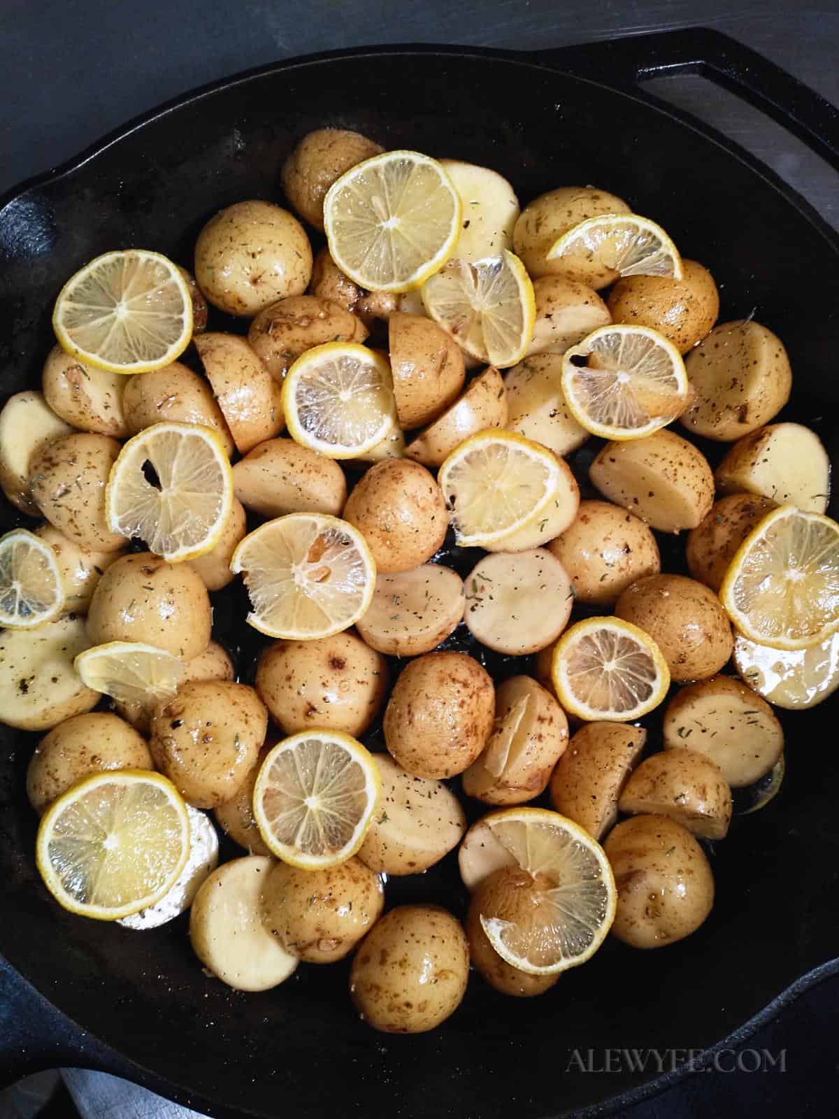 a cast iron skillet with thinly sliced lemon on halved new potatoes glistening with olive oil and herbs ready to roast