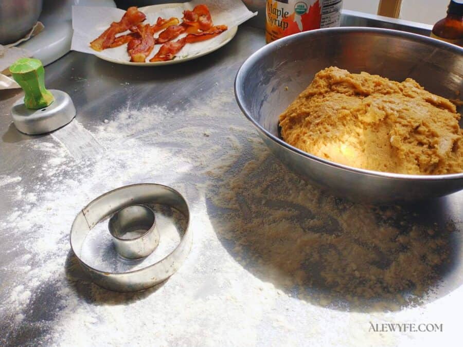 a bowl of maple bacon protein doughnut dough ready to roll out on a floured work surface with cutters. a plate of bacon is in the background.