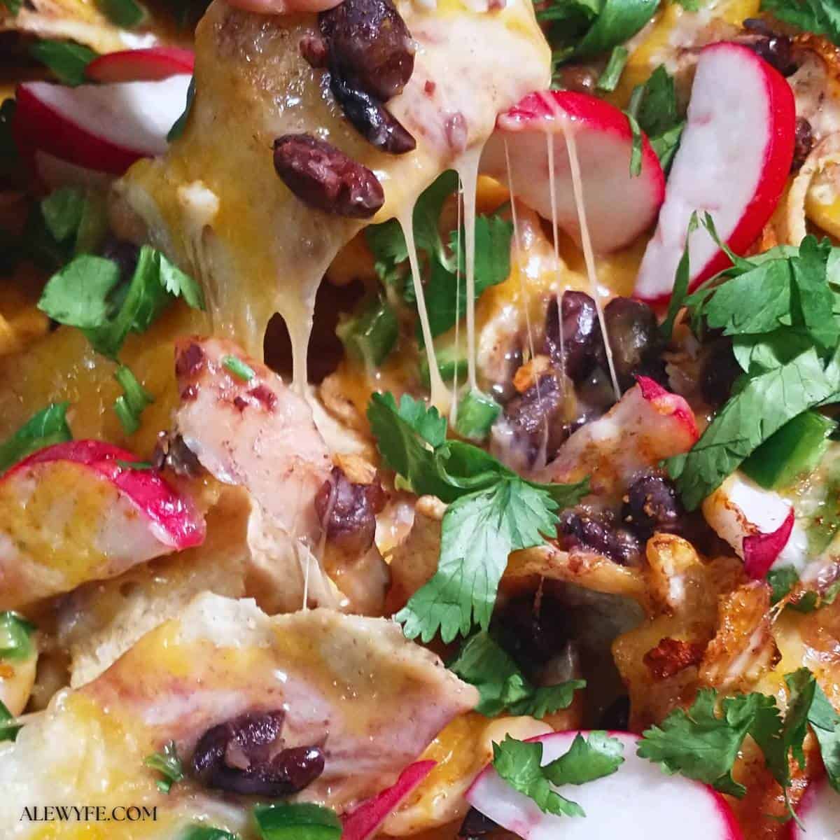 close-up view of nachos piled with melted cheese, cilantro, black beans, chicken, and vegetables, with stretchy melted cheese