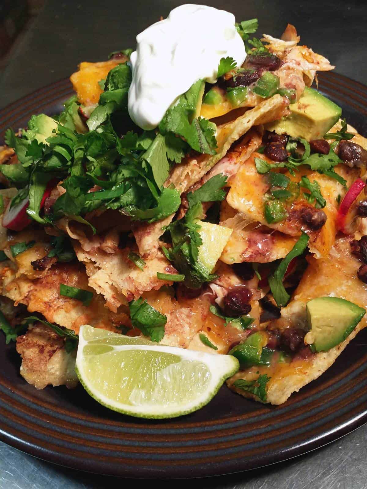 a brown ceramic plate heaped with a tall pile of black bean and cheese nachos, garnished with veggies, cilantro, lime, and sour cream