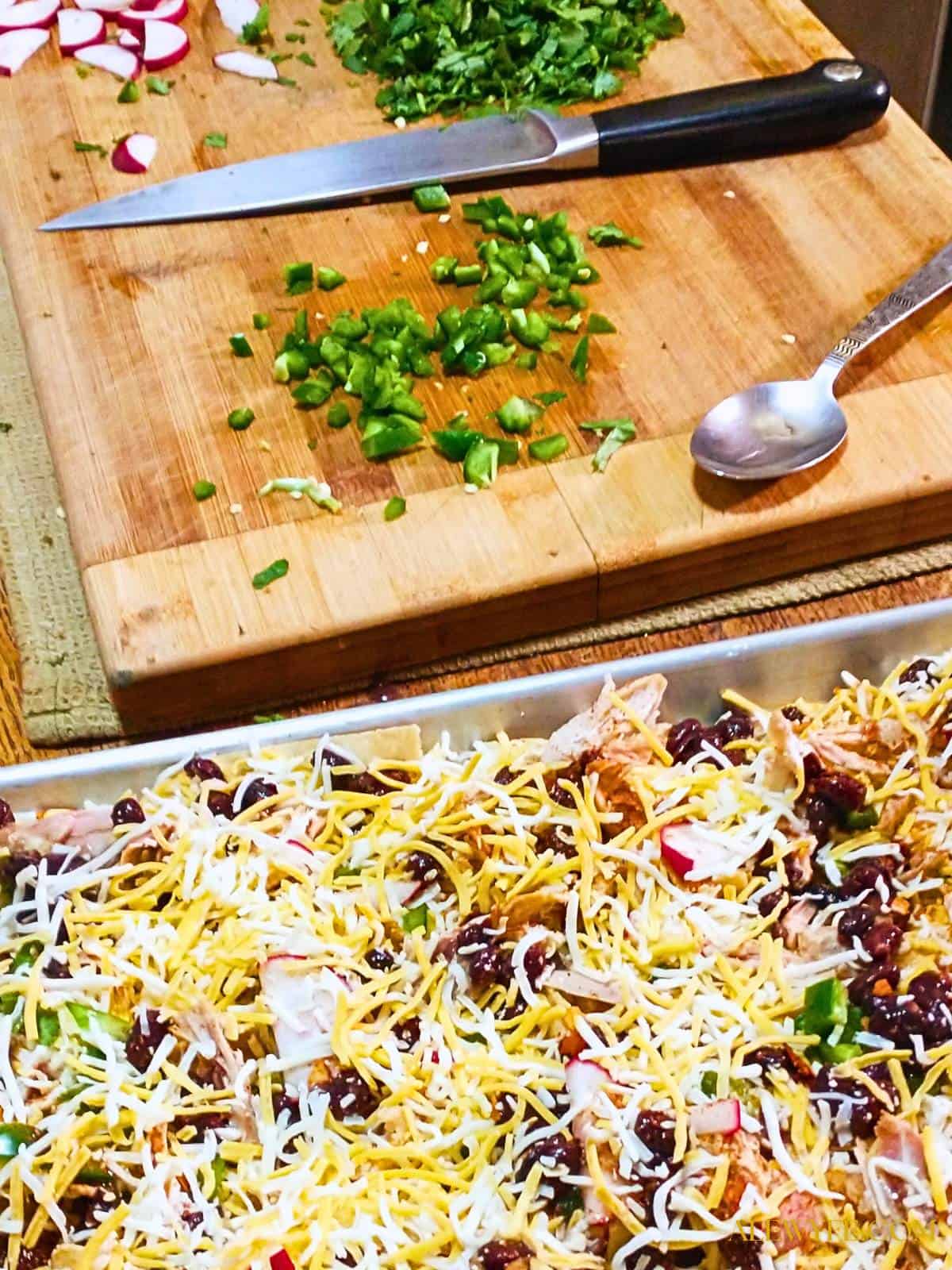 a wooden cutting board with chopped vegetables and a chef's knife is in the background and a sheet pan of nachos layered with cheese, beans, meat, and vegetables and ready to bake
