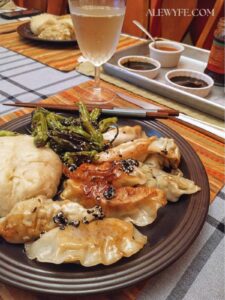a dinner table set with plates piled with dumplings, pot stickers, a steamed bao bun, and shishito peppers. A cold glass of white wine is in front of a tray of several dipping sauces and a spoon. 