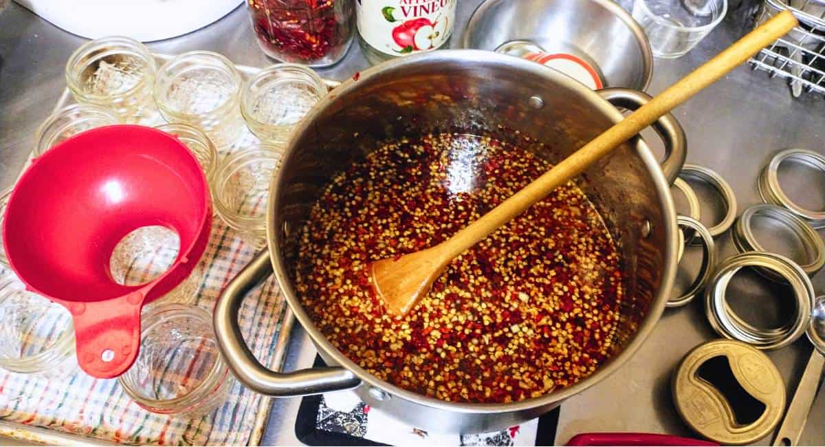 a pot of sweet chili-garlic sauce ready to put into mason jars. The jars are on a checkered tea towel, and an assortment of canning rings and a funnel are nearby.