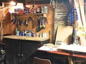 a wooden shelf with rows of hanging tools above a workbench in a basement workshop. A miter saw and bar clamps are on the right.
