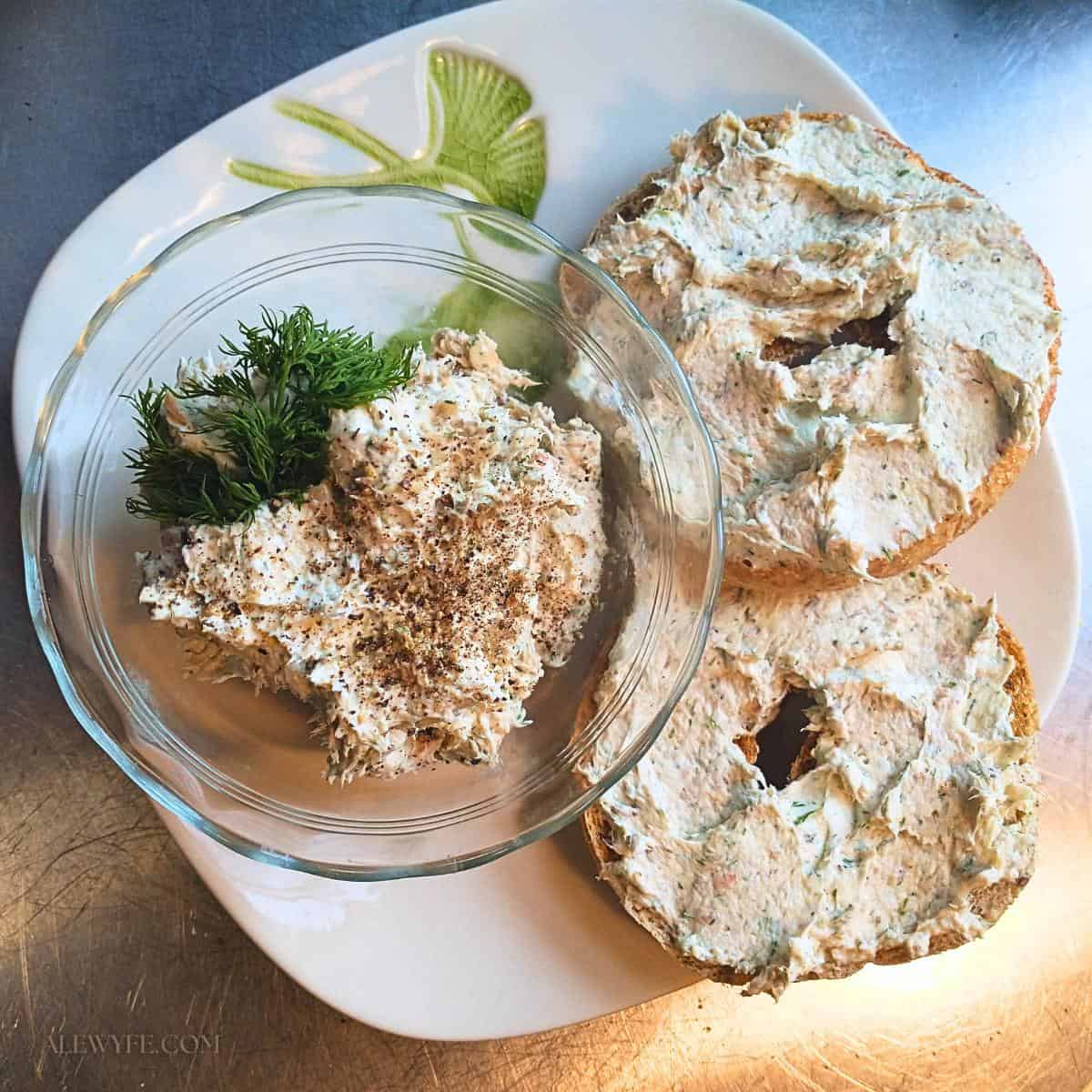 a bowl of smoked trout spread garnished with fresh dill, on a plate with two whole grain bagels.