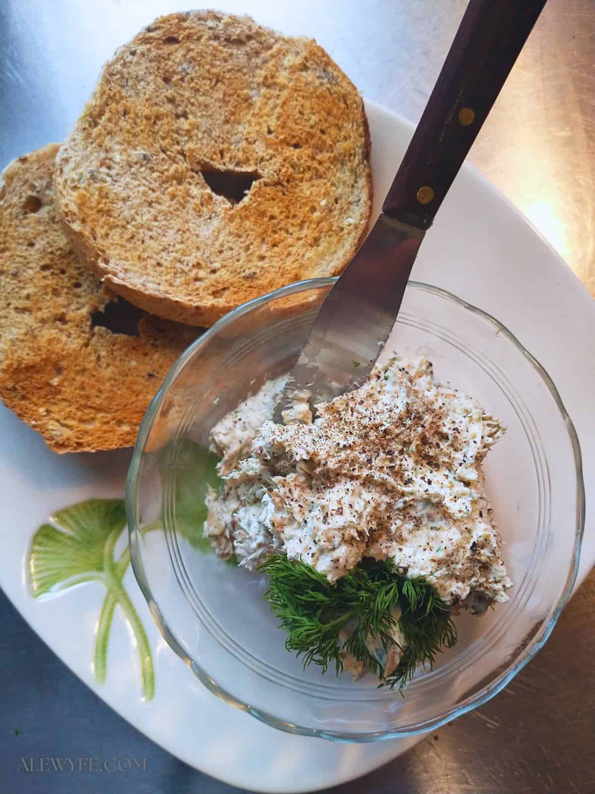 a sliced toasted whole wheat bagel on a plate with a ginko-leaf pattern. A glass bowl of smoked trout dip with a sprig of dill and fresh black pepper is on the plate, along with a wood-handled spreader knife.