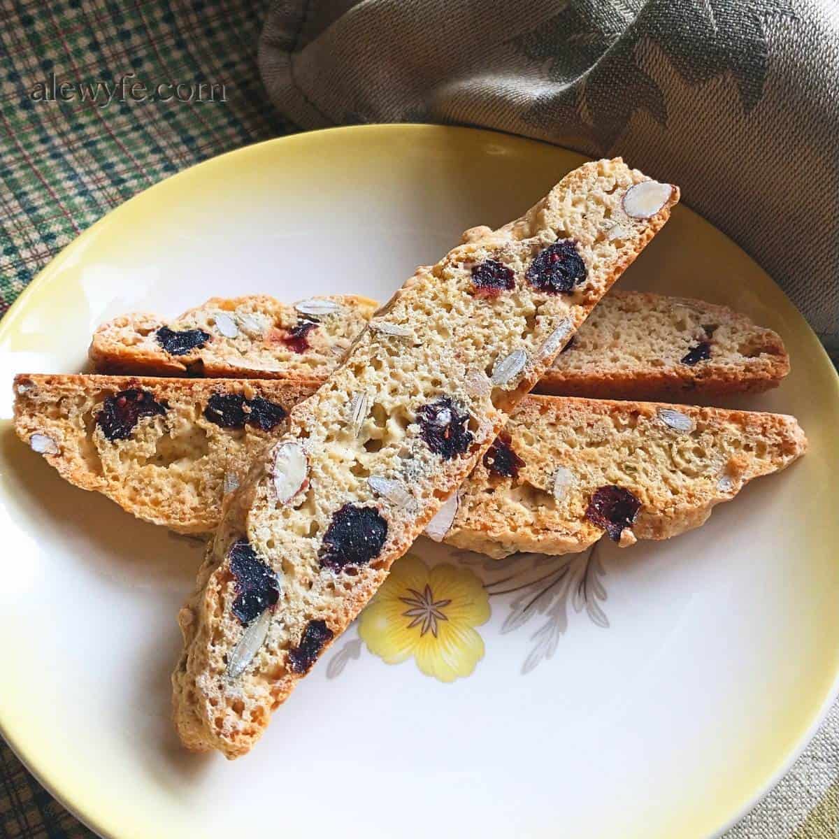 Three biscotti studded with almonds, pepitas, and dried cranberries. The cookies are on a vintage mid-century ceramic plate with a yellow rim and a flower.