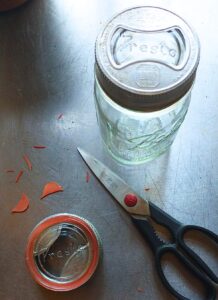 Two vintage glass mason jar lids, a jar, and trimming the tab from a Weck canning jar seal to fit inside the jar ring with a pair of kitchen shears.