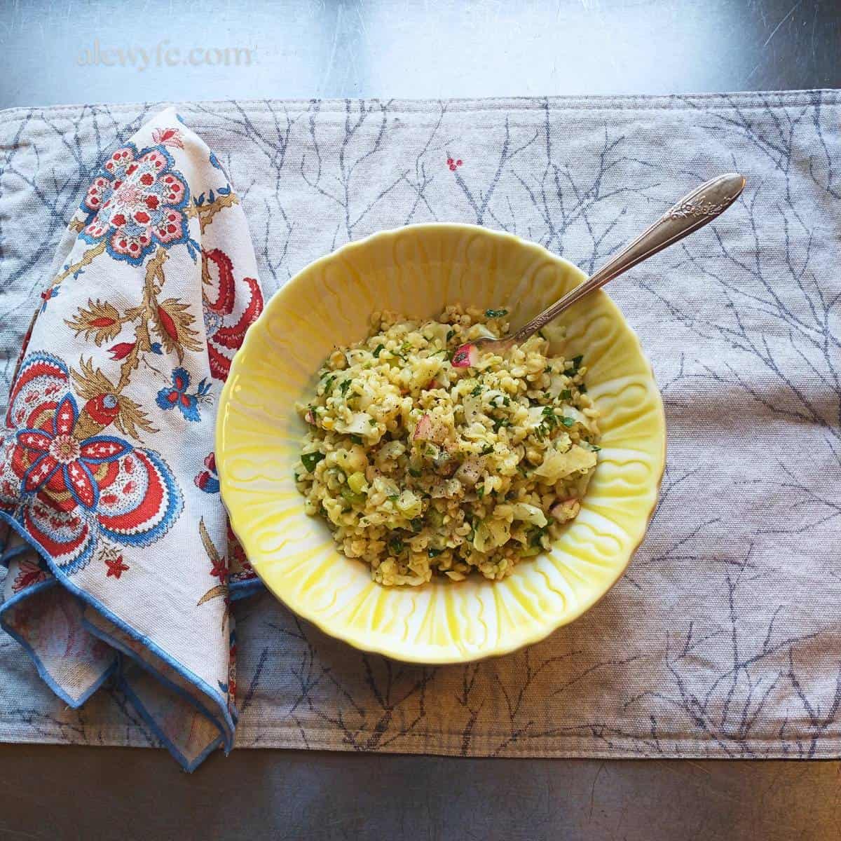 A bowl of winter tabbouleh salad on a placemat with a winter tree print, and a brightly colored paisley fabric napkin.