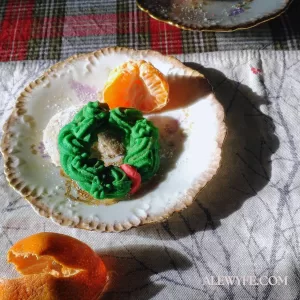 homemade spritz yule cookies with a clementine on vintage limoges china plate with gilt trim staged on festive table linens with winter branch print placemat and red, green, and black plaid tablecloth.