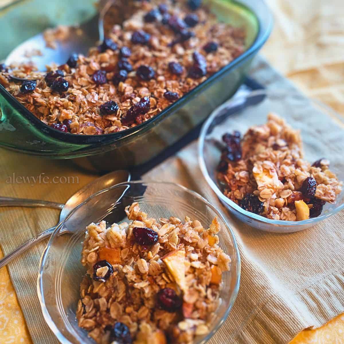 a green glass pyrex dish with baked apple cranberry walnut oatmeal, with two clear glass bowls of baked oats and silver spoons in the foreground.